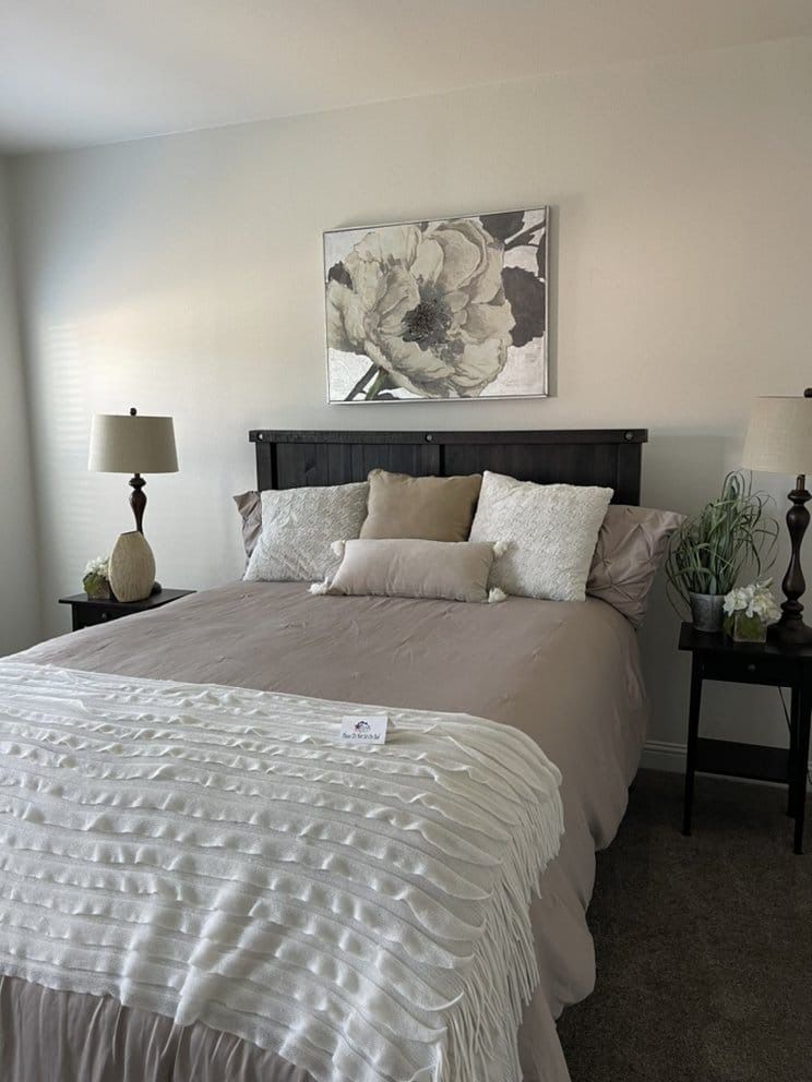 Bedroom with a bed, nightstands, lamps, and floral artwork. Taupe and white color scheme.