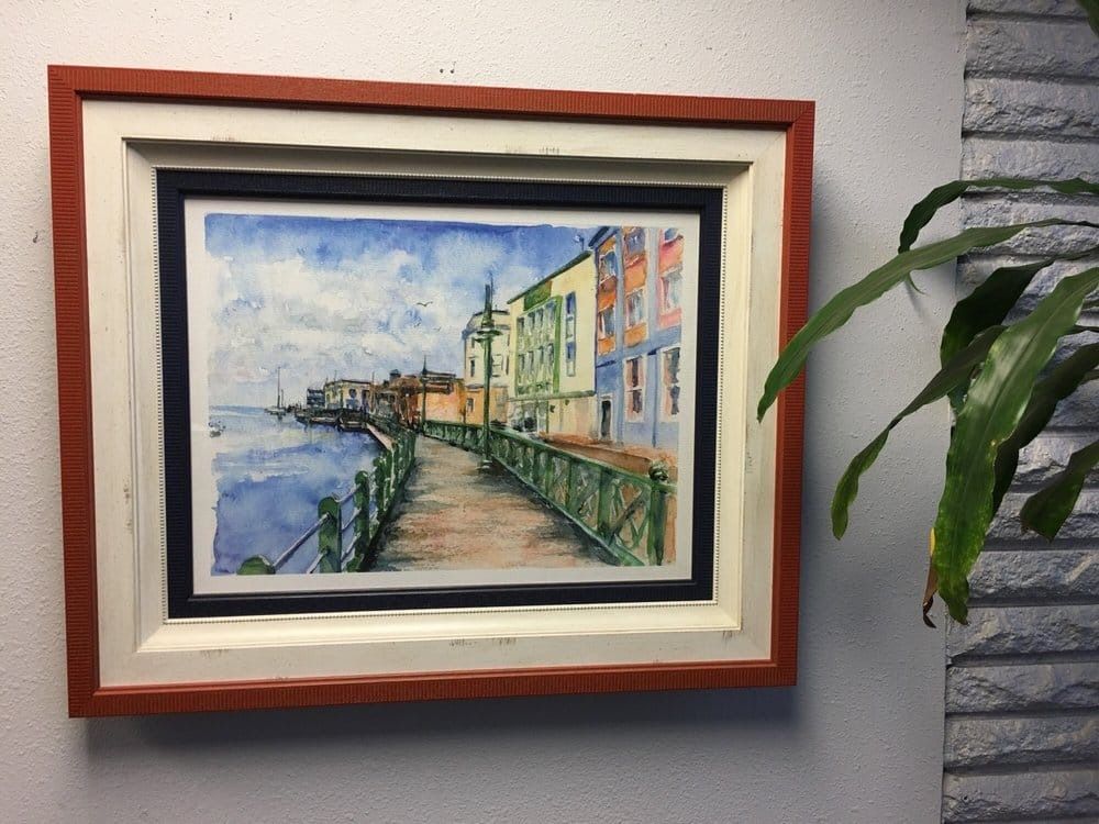Watercolor painting of a waterfront path with colorful buildings, framed and hanging on a wall next to a plant.