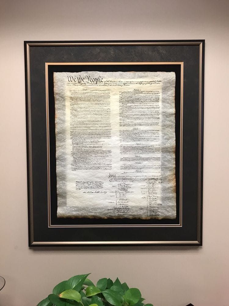 Framed document of the U.S. Constitution, mounted on a wall. The paper is aged, with black and gold frame.