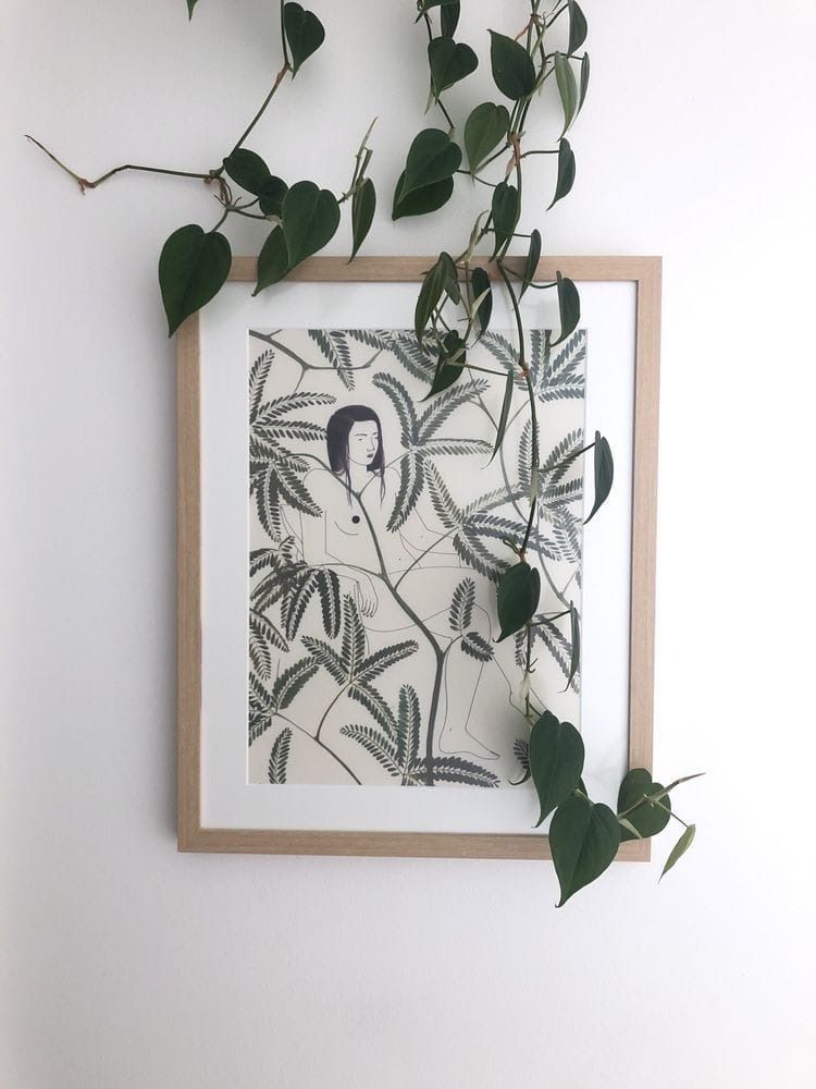 Framed art with a person amid foliage, hanging on a white wall; green vine draping over the frame.