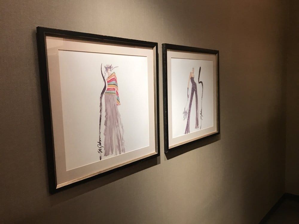Two framed fashion sketches on a beige wall; one of a dress with colorful bodice, the other of pants.