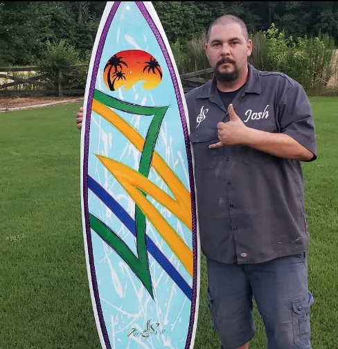 Man standing beside a colorful surfboard with a sunset and palm tree design on a lawn