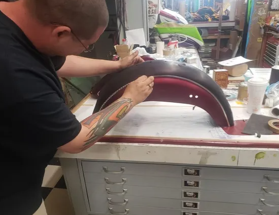 Person painting a maroon curved car part at a workbench in a workshop