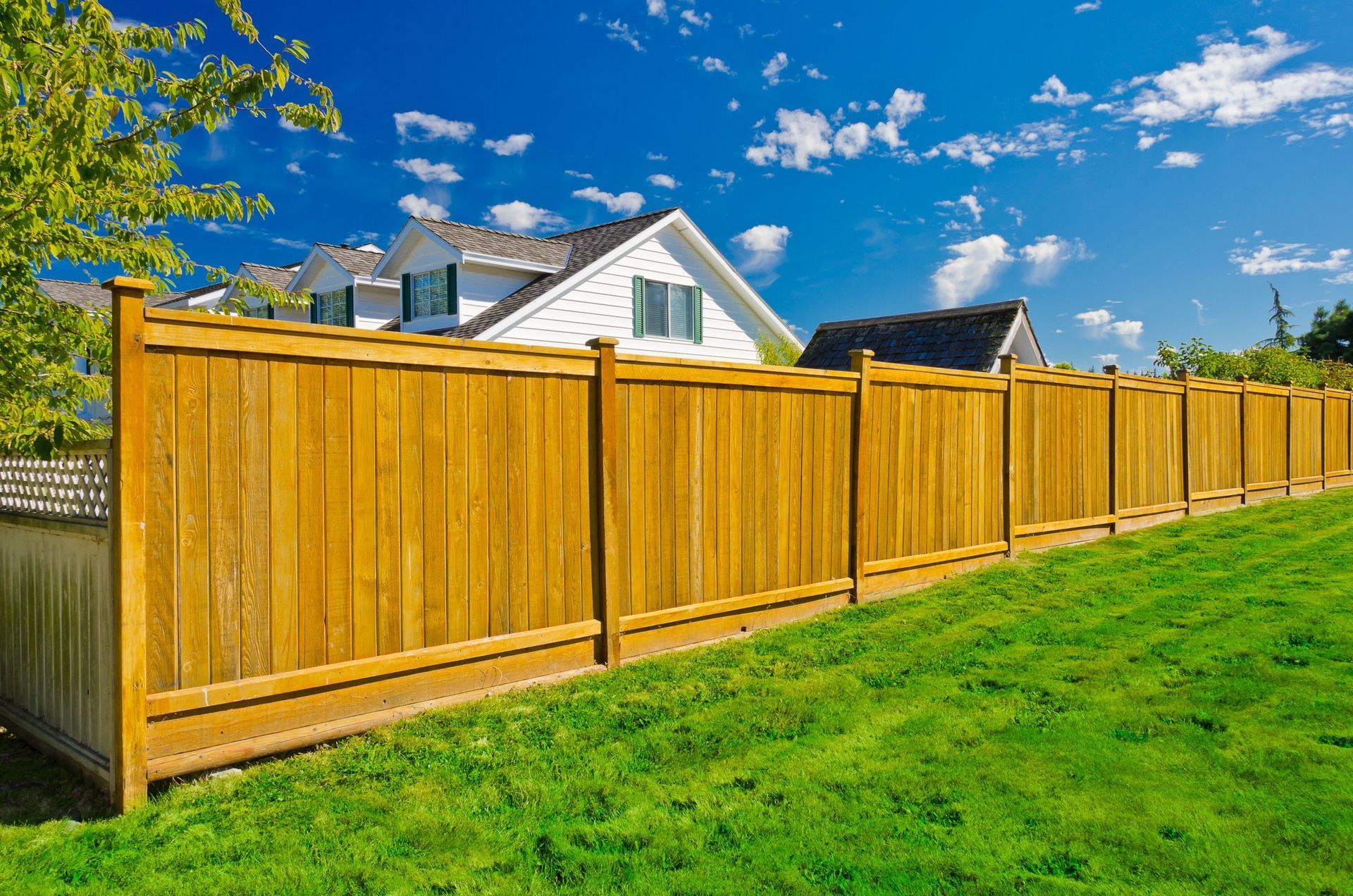 Wooden fence in front of a white house on a sunny day with green grass and blue sky.