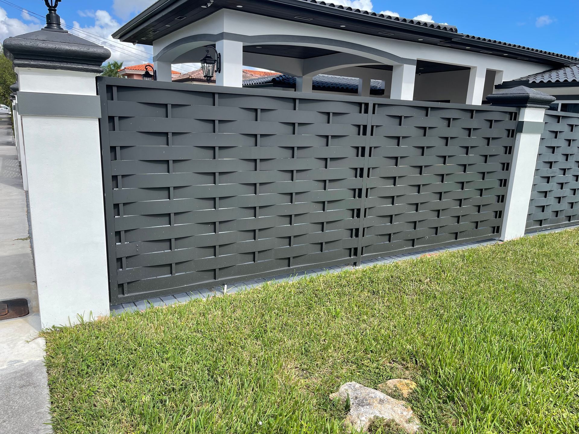 Gray woven-pattern fence with white pillars, surrounding a house. Green grass in foreground.
