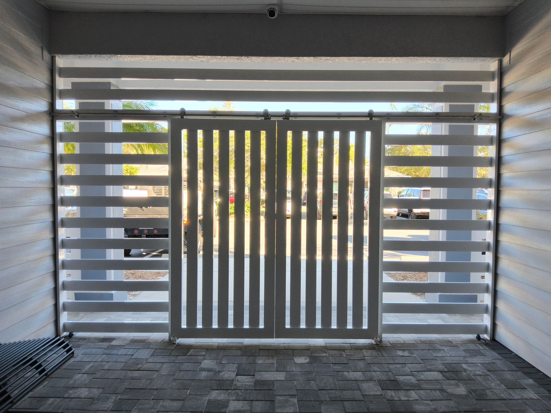 White slatted gates with vertical bars, surrounded by horizontal slatted blinds, set in a brick-paved area.