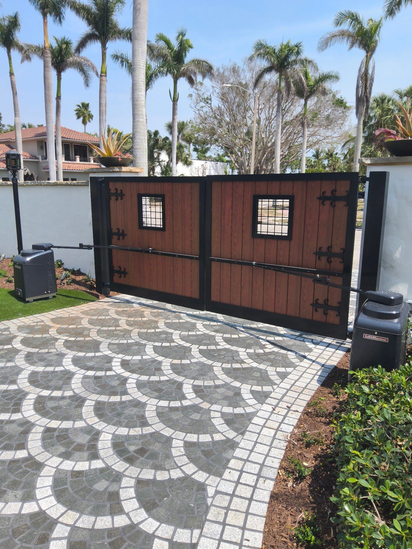 Brown wooden driveway gates with iron hardware, opening onto a stone-patterned driveway. Palm trees and a house are in the background.