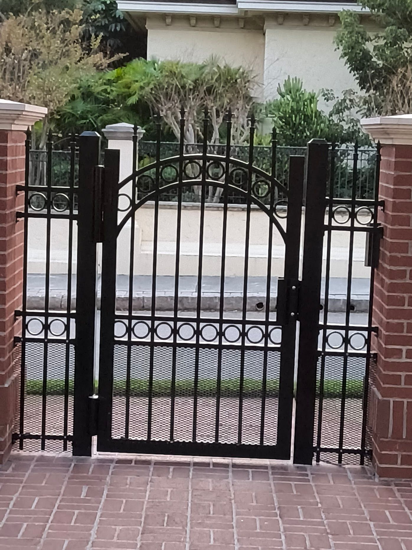 Black metal gate with arched top, between brick pillars. A brick walkway leads to the gate.