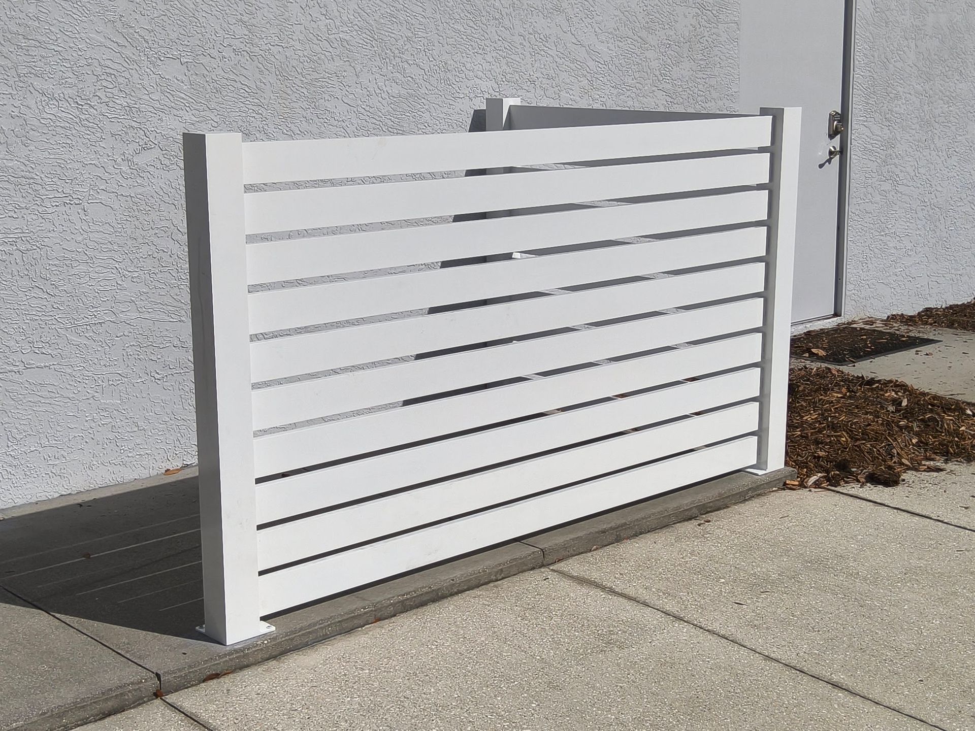 White horizontal slat fence against a light-colored building, on a concrete sidewalk.