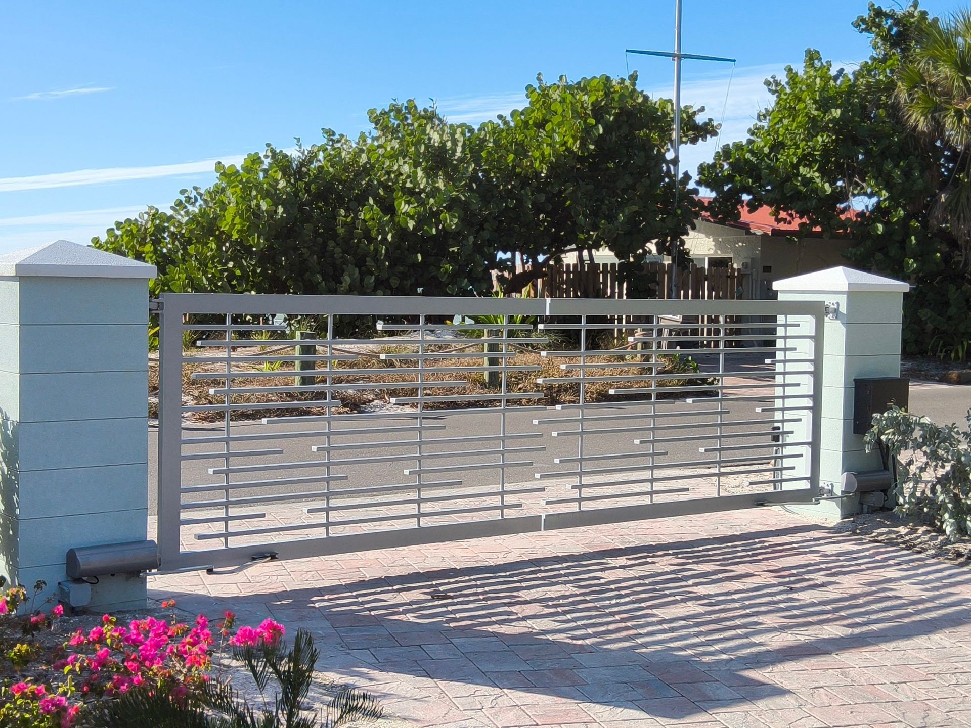 Gray metal gate with horizontal bars between light blue pillars. Green trees and blue sky background.