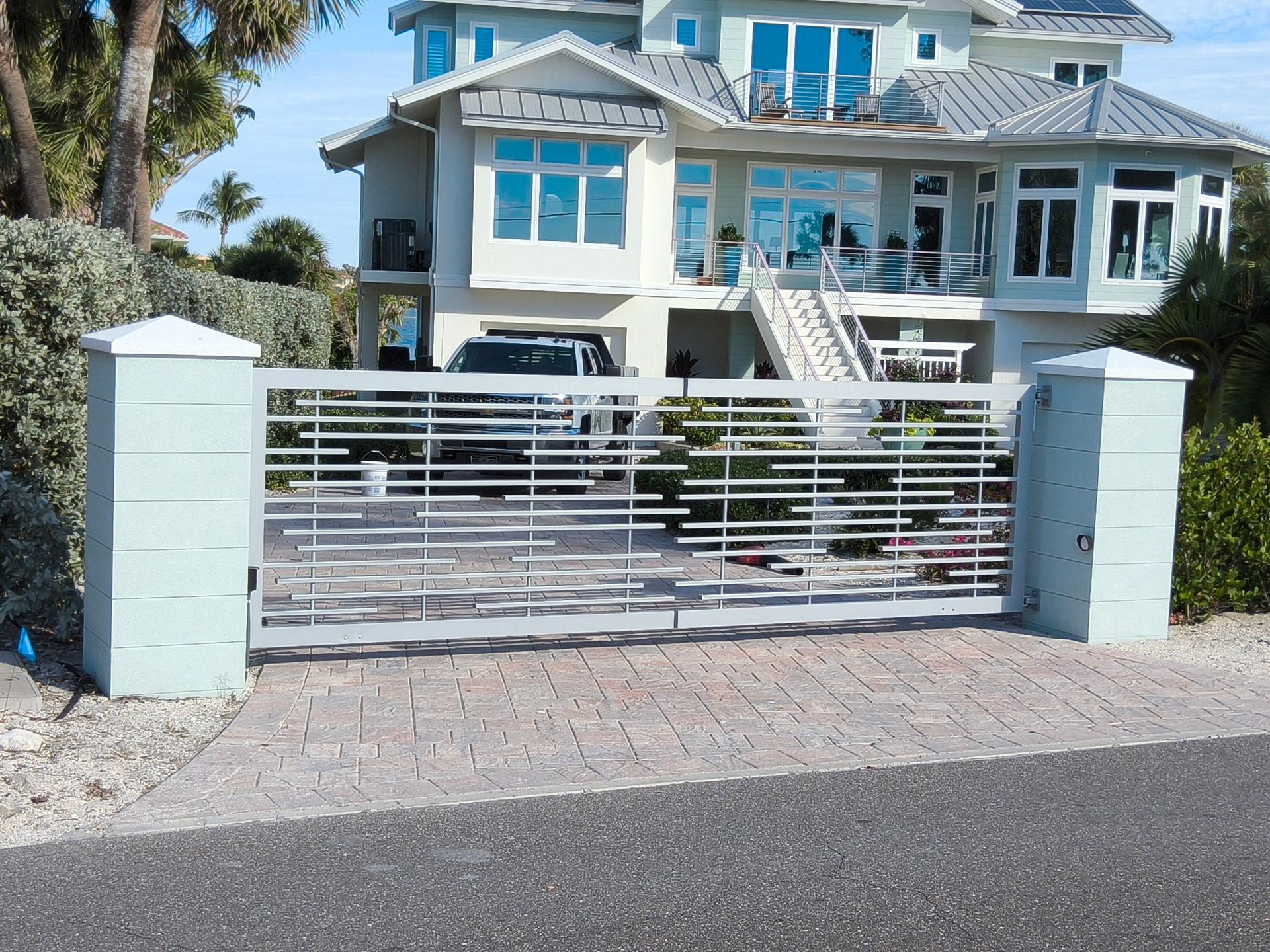 A light gray metal gate leading to a large turquoise house with a gray roof and a brick driveway.