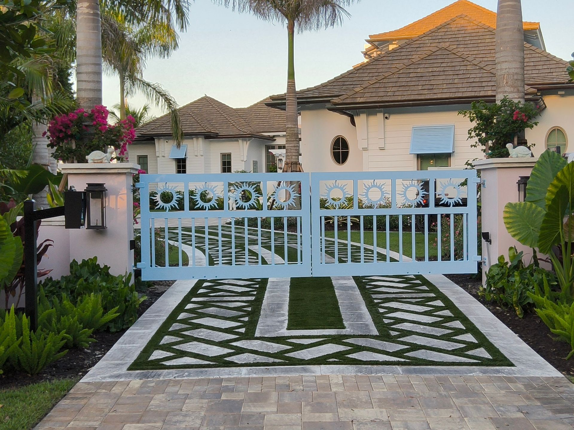 Light blue gate in front of a house with a patterned driveway, lush landscaping, and pink pillars.