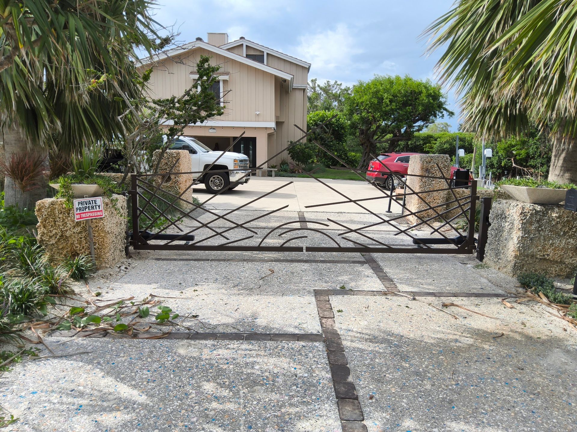 Wrought iron gate in front of a light-colored house.  Driveway with parked truck and car, palm trees.