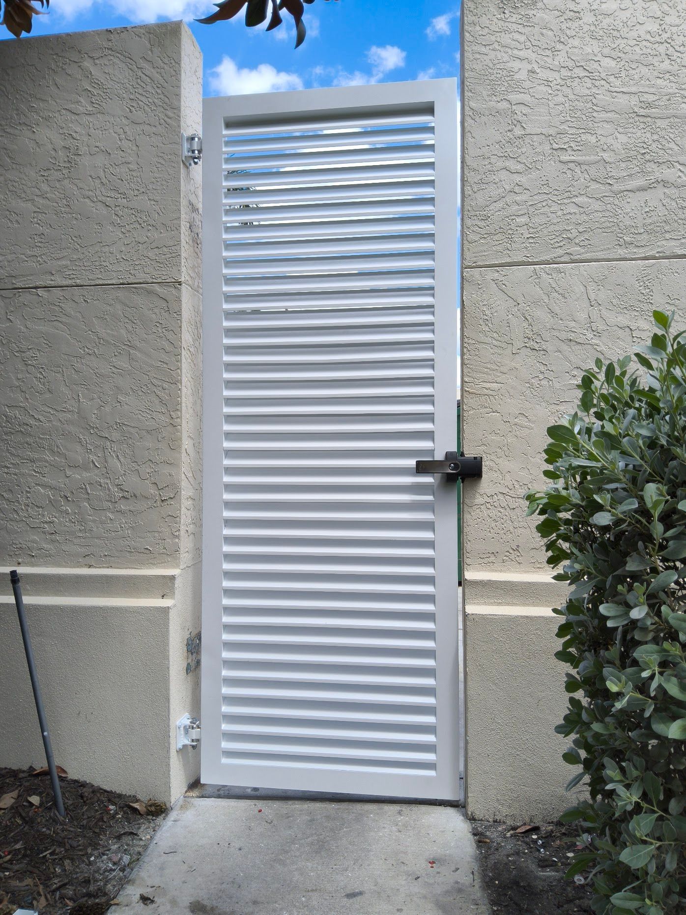 White gate with horizontal slats, between two stucco walls, a latch, and a small bush.