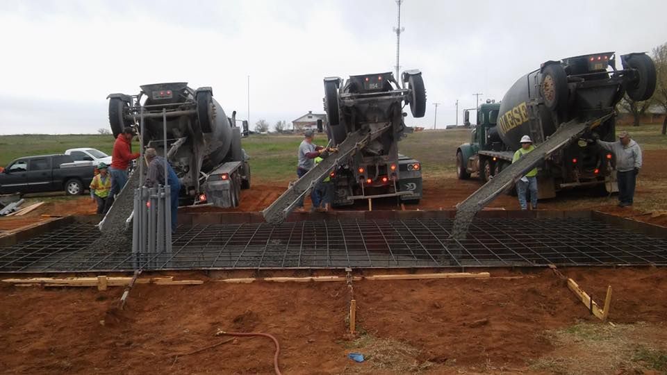 Three cement trucks pour concrete onto a rebar-reinforced foundation on a dirt construction site.