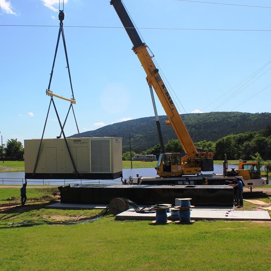 A yellow crane lifts a large industrial generator over a concrete base near a pond on a sunny day.