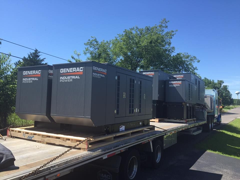Four grey Generac industrial power generators secured on a flatbed trailer outdoors on a sunny day.