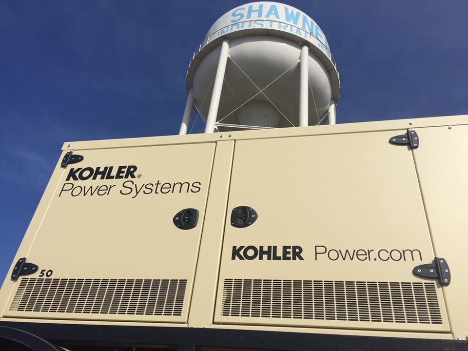 A tan Kohler Power Systems generator sits outdoors beneath a large, light-colored water tower against a blue sky.