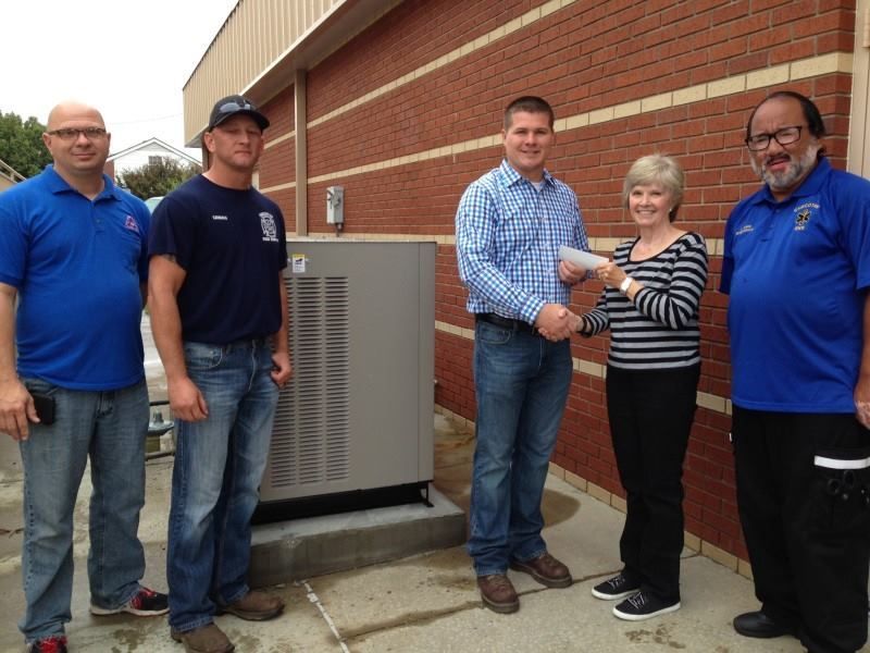 Four people stand outside by a large outdoor HVAC unit, with one person handing a check to another.