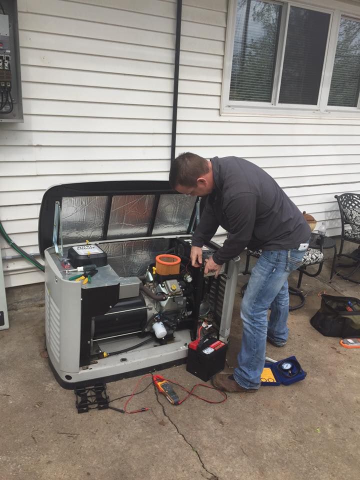 A technician in a gray sweatshirt repairs an open standby generator outside a house, using a multimeter on the ground.