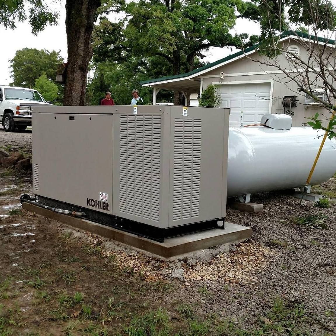 A tan Kohler backup generator and a white propane tank sit on a concrete pad outside a house in a wooded area.