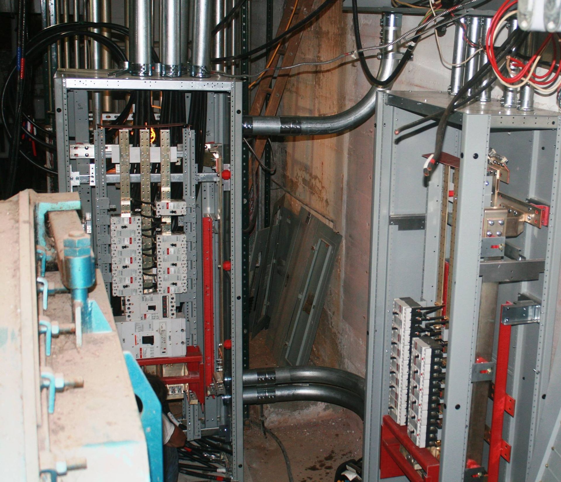 Open electrical panels with exposed metal bussing, circuit breakers, and conduit wiring in an industrial setting.