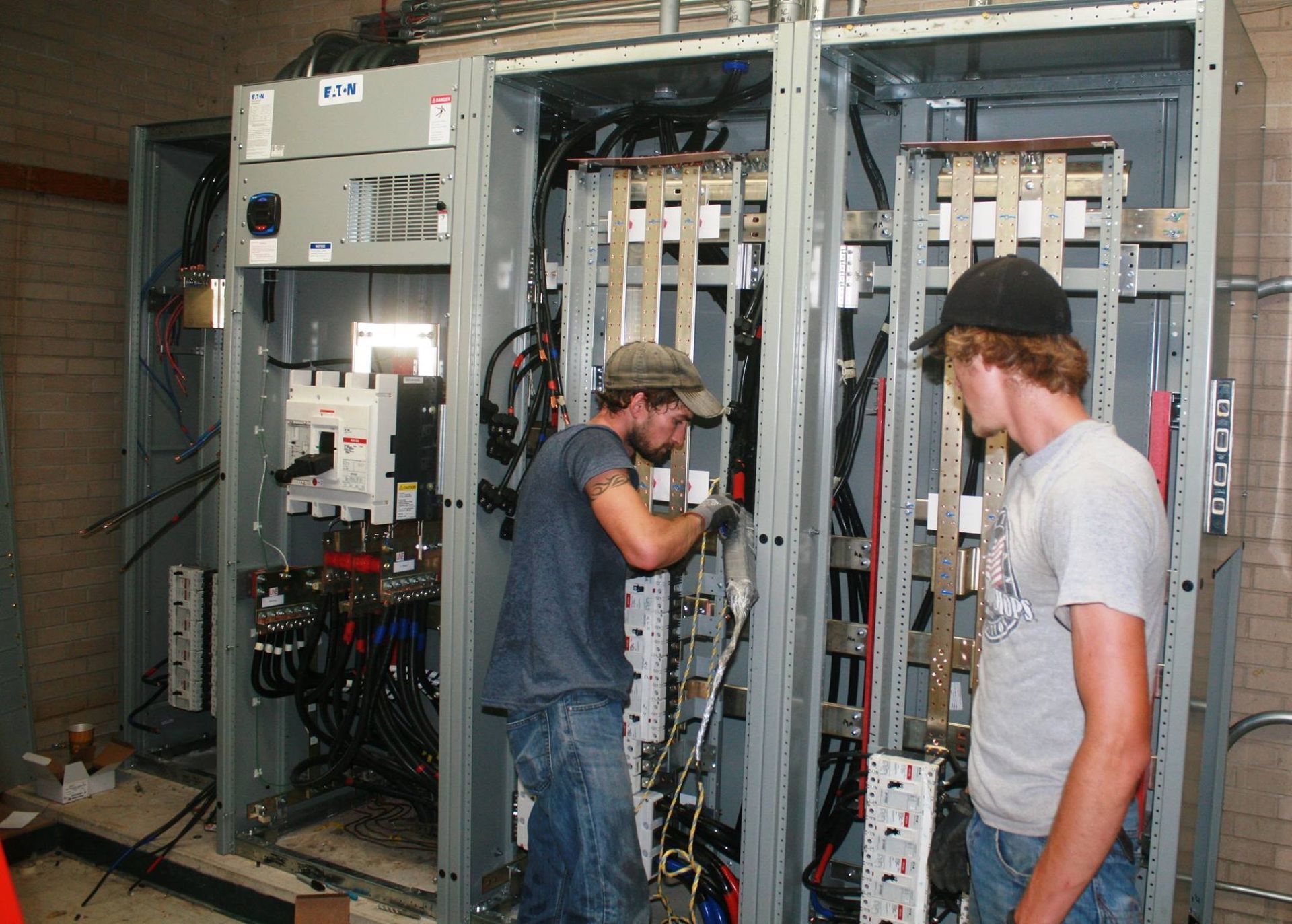 Two workers in casual clothing install wiring and components inside a large, open industrial electrical switchgear cabinet.