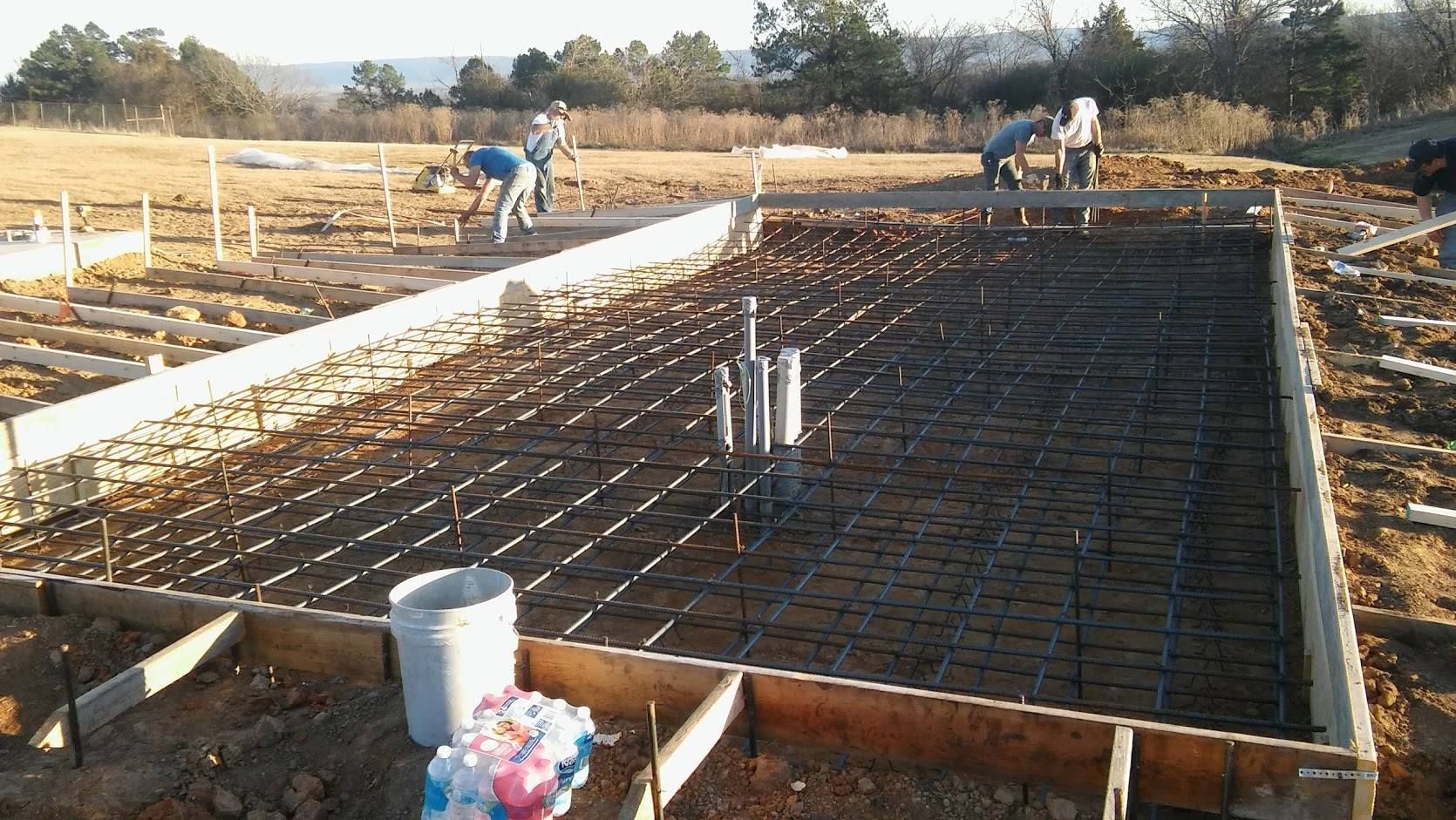 Workers prepare a construction site with a wooden-framed foundation and a steel rebar grid before pouring concrete.