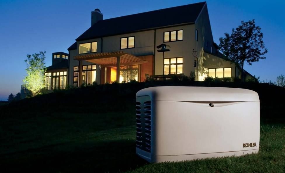 A white Kohler home standby generator sits on a grassy lawn in front of a modern house illuminated at dusk.