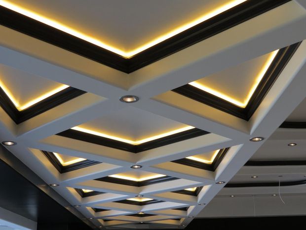 A modern coffered ceiling with white geometric beams, black recessed edges, warm accent lighting, and small spotlights.