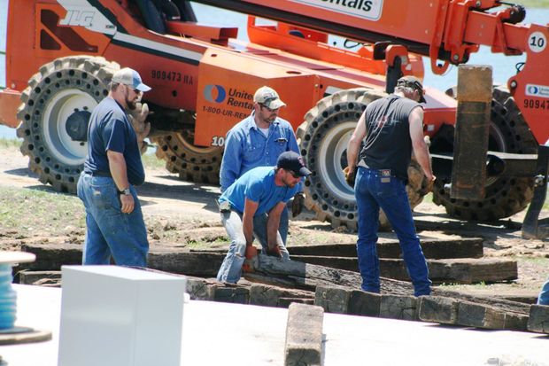 Four workers in casual work clothes lift heavy wooden beams on a job site with an orange construction telehandler.