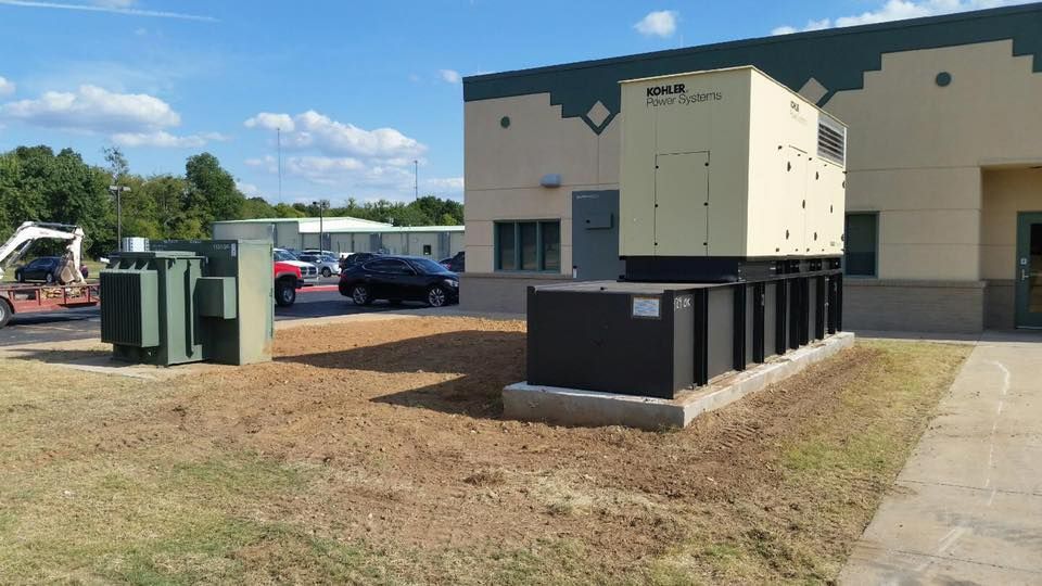 A large industrial power generator and electrical transformer sit on a grass lot outside a beige commercial building.