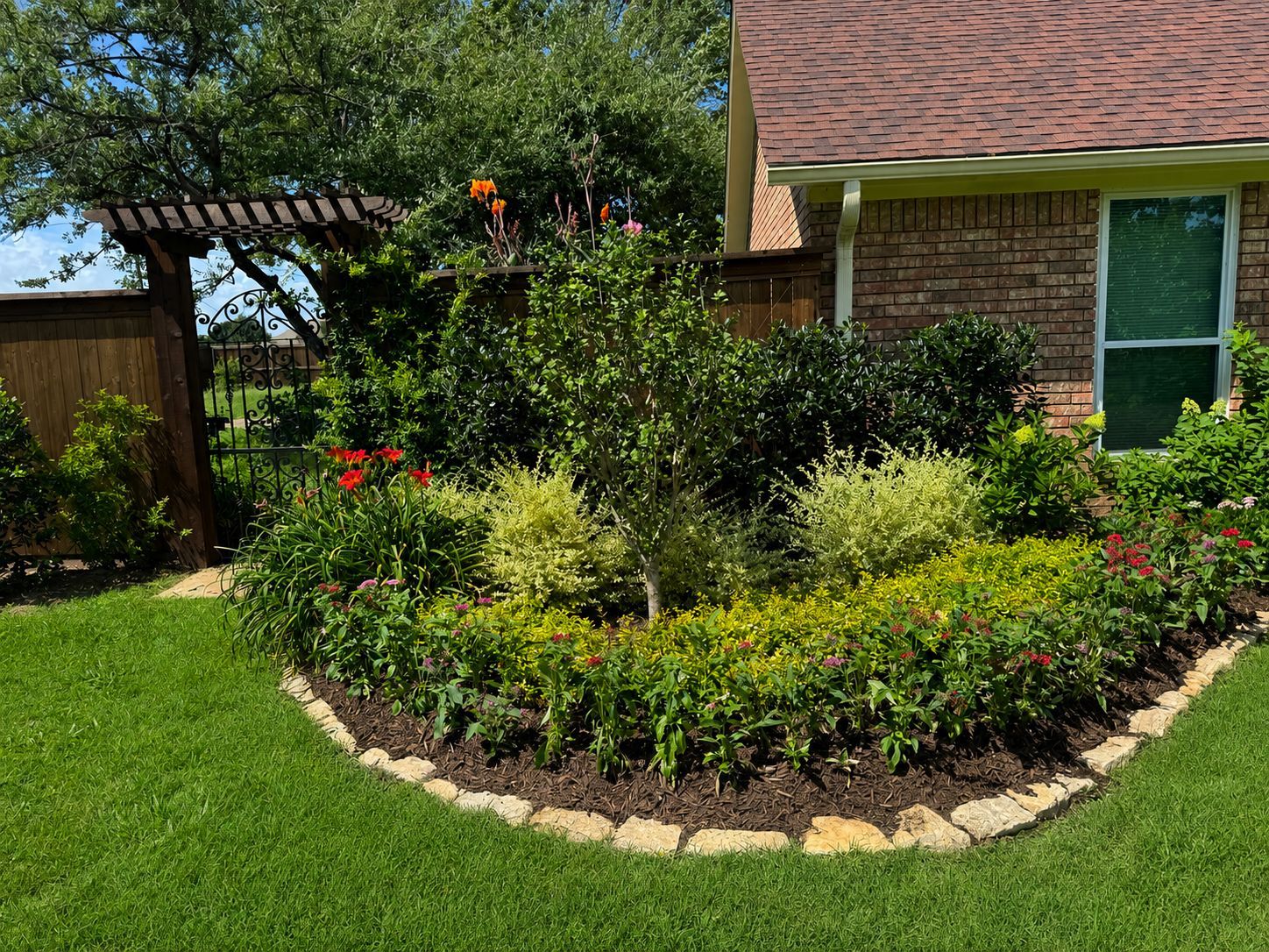 Landscaped garden bed with red flowers, shrubs, and stone edging beside a brick house