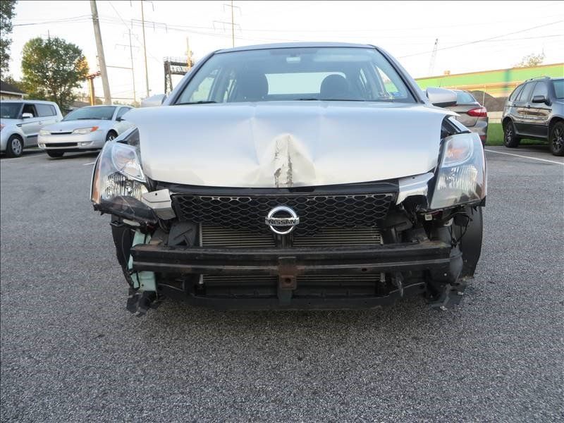 A silver nissan car with a damaged front end is parked in a parking lot