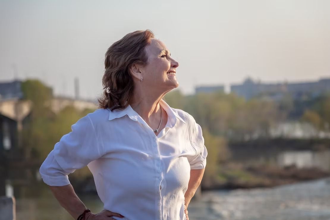 Woman in a white shirt standing outdoors, smiling up toward the sky by the water at sunset