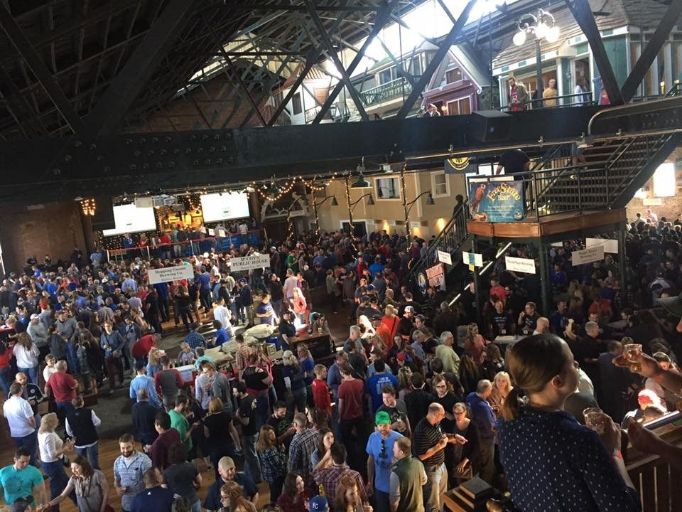 Large crowd of people gathered indoors, possibly for an event. Many are standing, some holding signs, in a large open space with an industrial aesthetic.