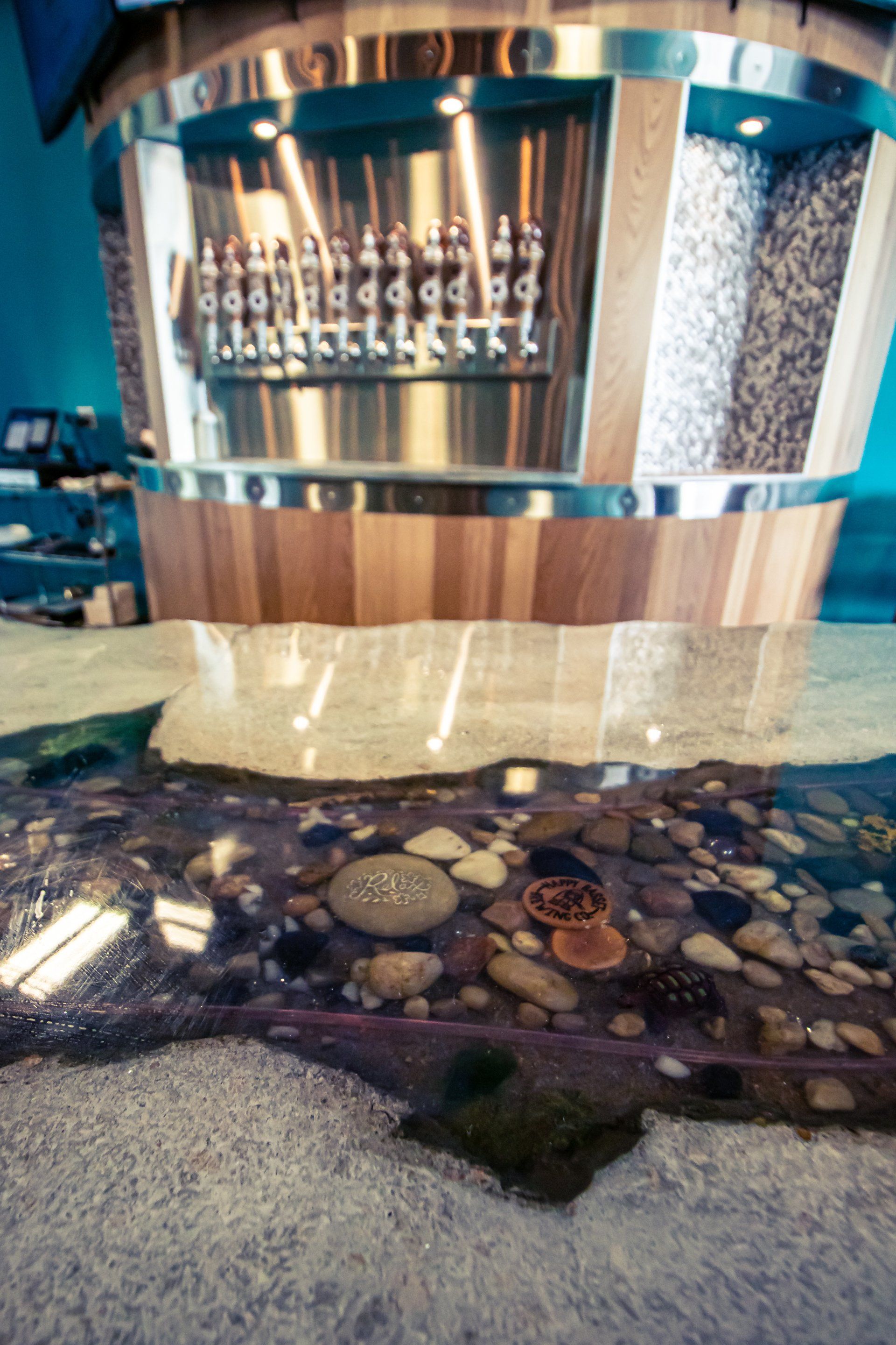 A beer tap counter with a river feature. The counter is stainless steel and wood, with taps in a row. The floor resembles a rocky stream.