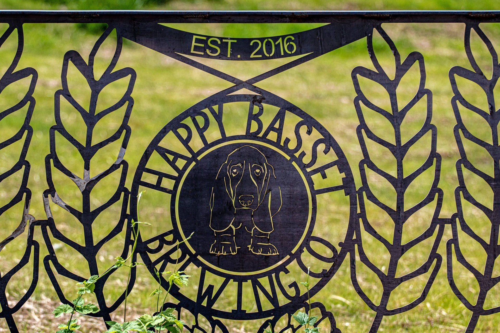 Black metal sign for Happy Basset Brewing Co. with a dog silhouette, wheat stalks, and the year 2016.