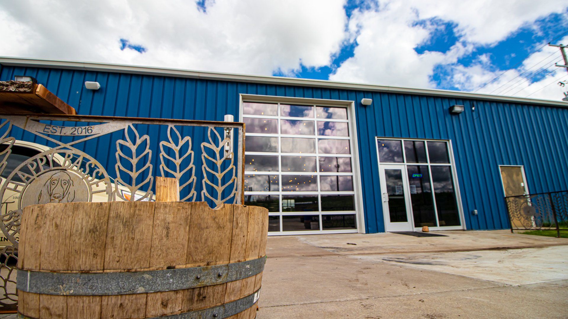 A blue building with a large glass door. A wooden barrel and metal sign with wheat art are in the foreground.