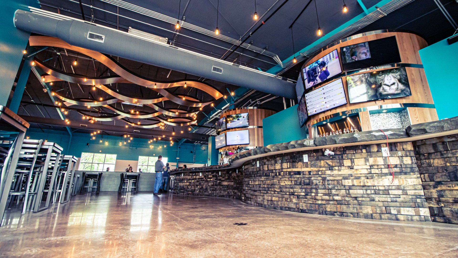 Interior of a modern sports bar with a stone bar, multiple TVs, and hanging lights. Teal walls and a glossy floor.