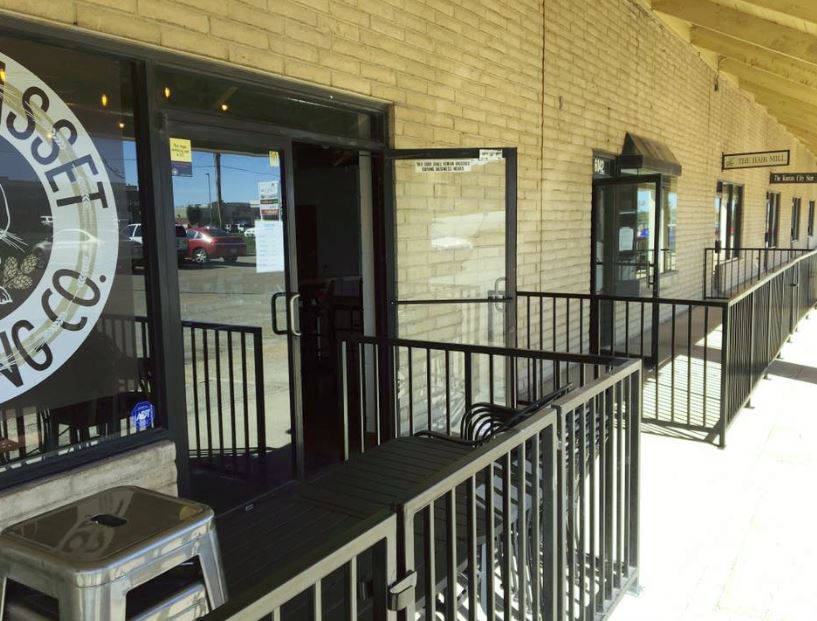 Exterior of a brewery with an open door, a small patio with railings, and a sidewalk under a canopy.