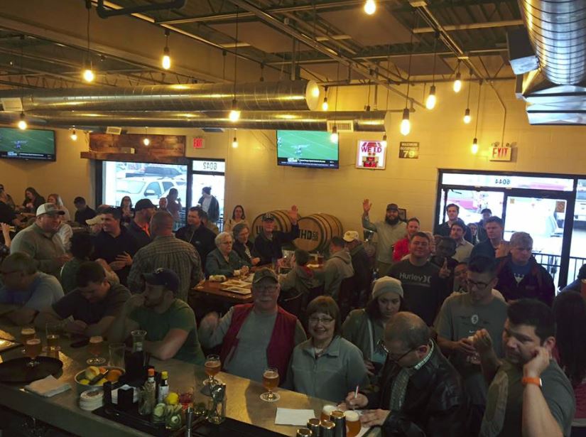 A crowded bar with people drinking, watching TVs, and socializing. Warm lighting, neutral tones, and a convivial atmosphere are evident.
