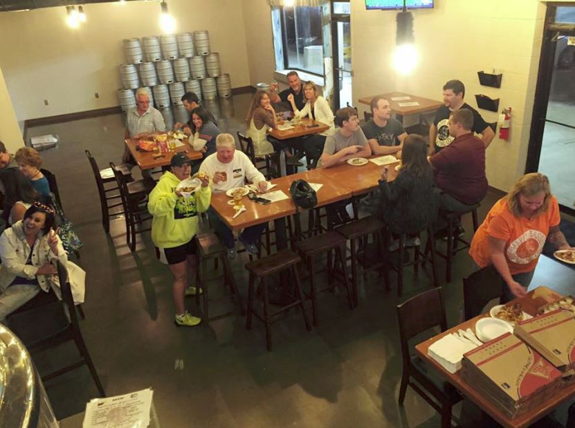 People gathered inside a brewery at wooden tables. Several are eating, one is taking food from pizza boxes, and kegs line the wall.