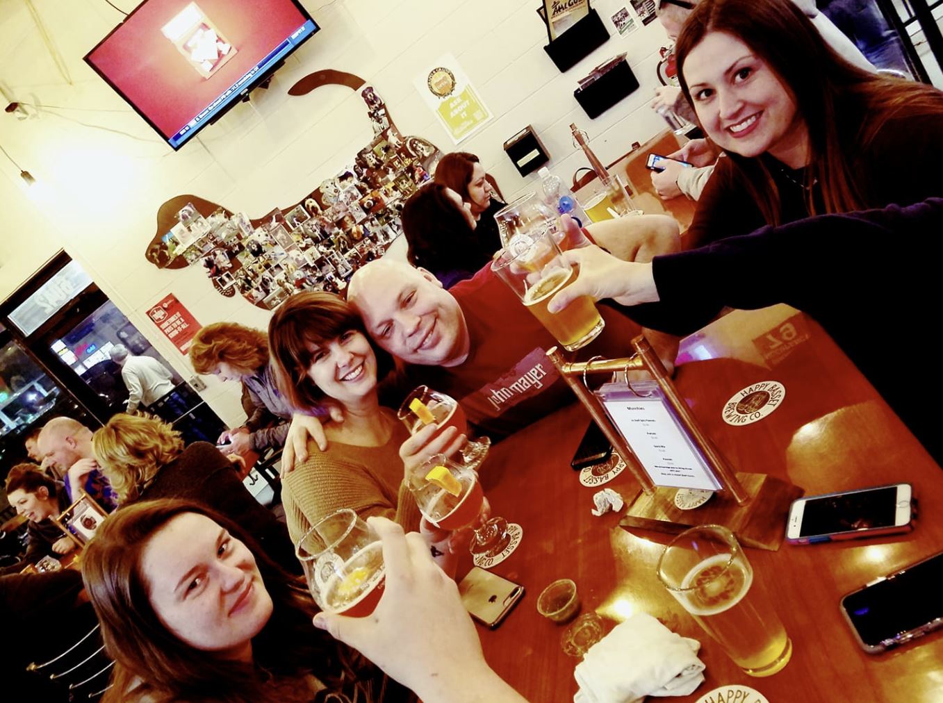 People toasting beers at a bar with a dachshund-shaped wall art, smiling faces, warm lighting, and a television.
