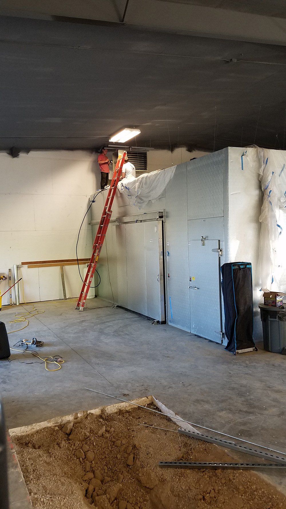 Interior of a room under construction. A tall metal structure with a closed door and ladder against it. Debris on the floor.