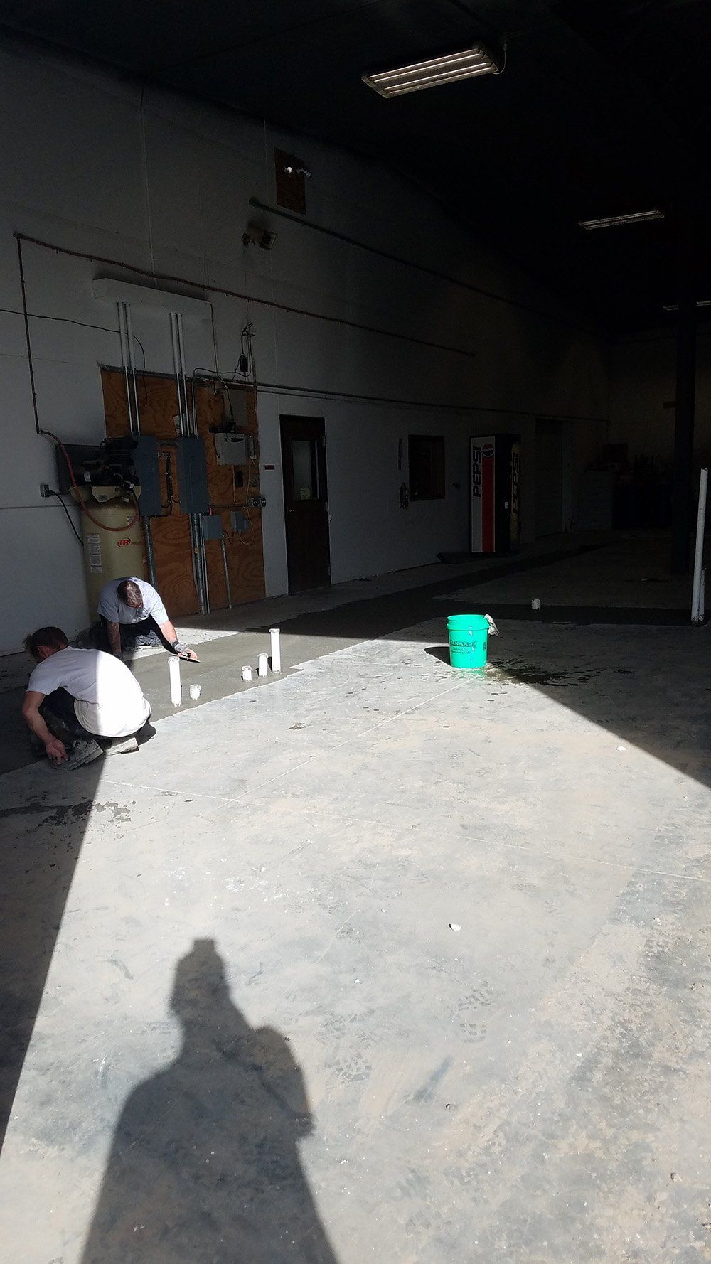 Two people working on plumbing inside a dim warehouse, near a green bucket. The warehouse floor is concrete.