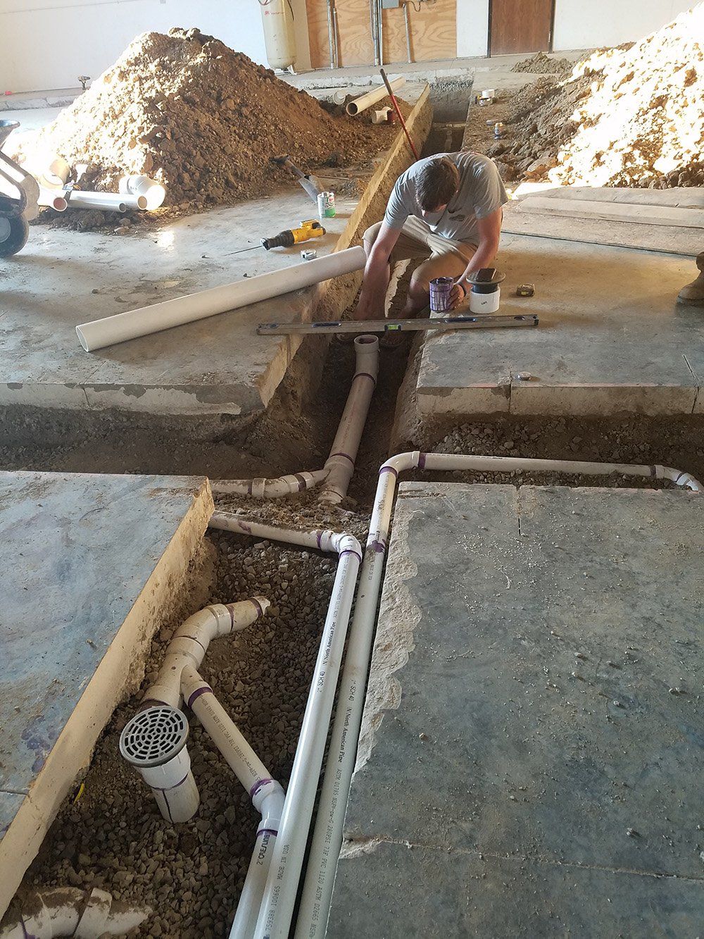 Plumber installing PVC pipes in concrete trenches. A pile of dirt is in the background.