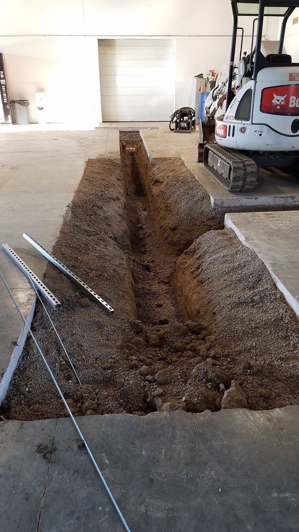 An interior shot shows an excavation in a concrete floor with a mini excavator and construction materials.
