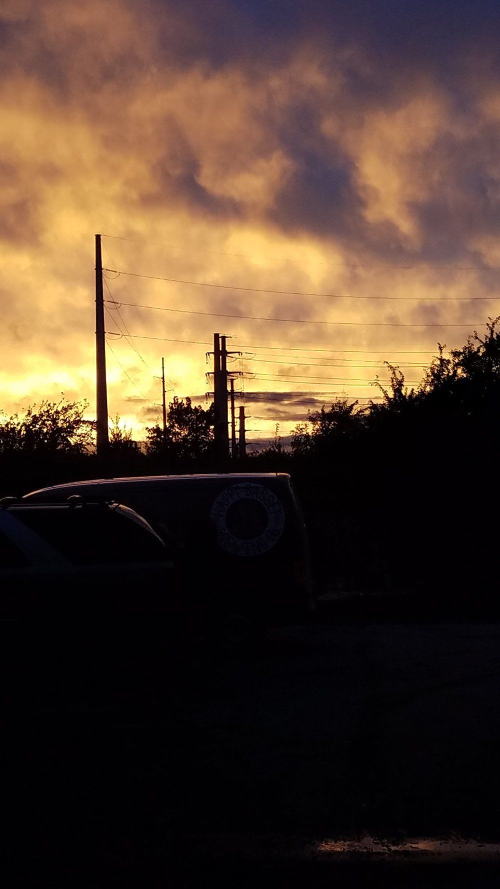 Sunset over silhouetted power lines, trees, and a parked vehicle, with a golden-yellow sky.