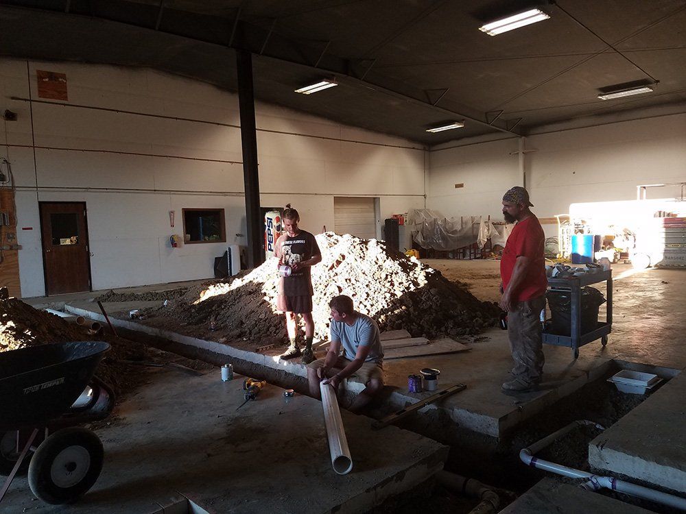 Three men work on plumbing in an indoor space. Dirt and a wheelbarrow are nearby. One man holds a pipe.